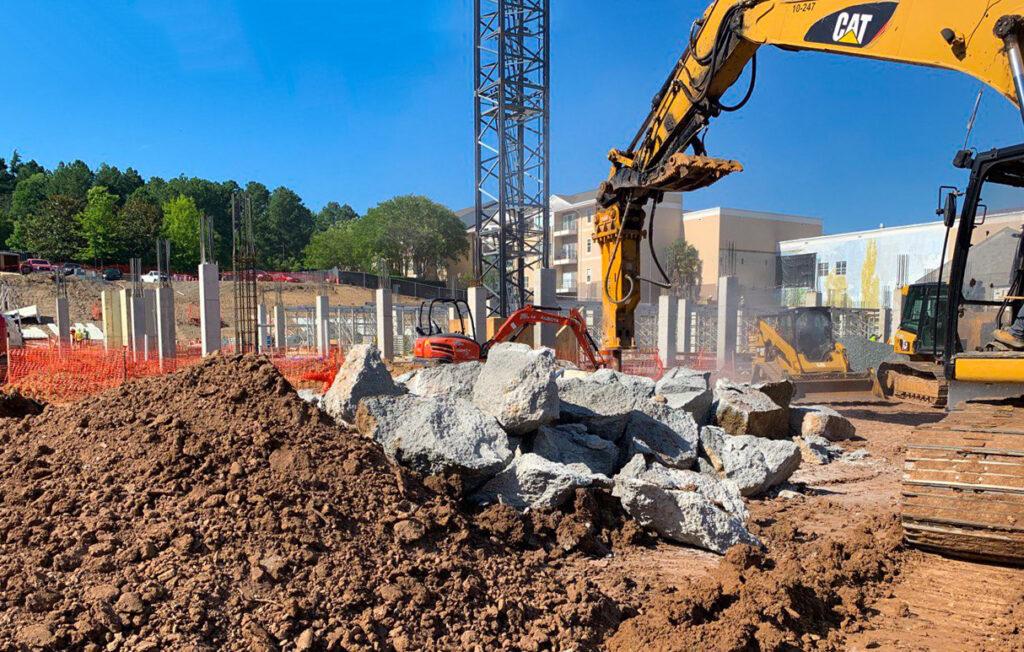 An East Coast Civil Group excavator with a hydraulic breaker attachment breaks up a concrete foundation for removal.